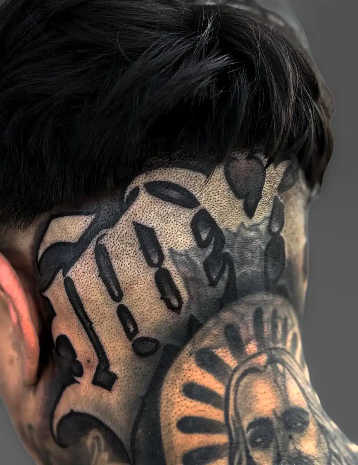 Black-and-gray blackletter tattoo across the side of the head and neck.